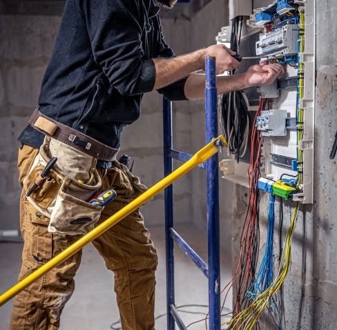 A male electrician works in a switchboard with an electrical connecting cable, connects the equipment with tools.